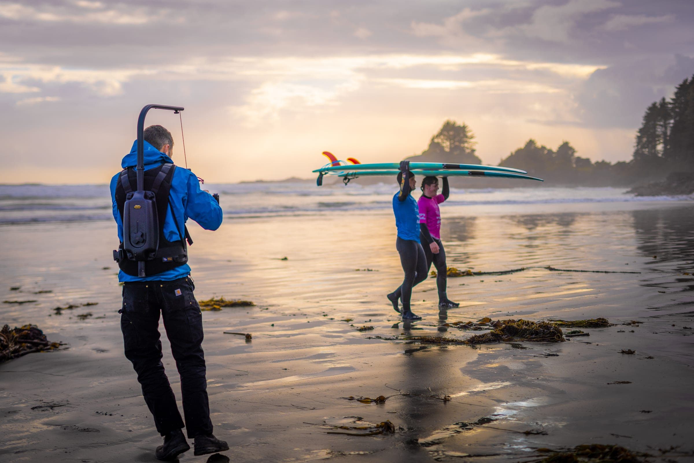 Two surfers carry a longboard across a wet beach at sunset while a camera operator films them from behind.